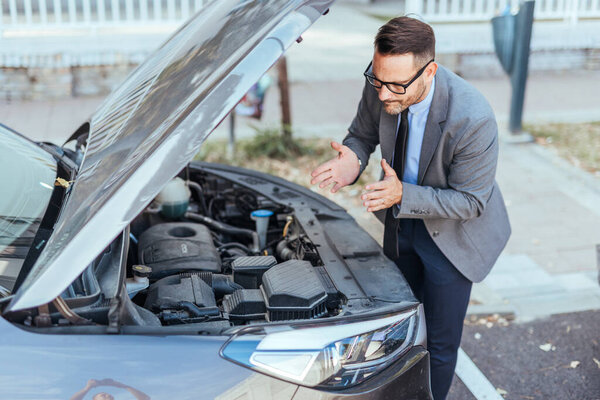 A business professional in formal attire checking under the hood of a car parked on the street, emphasizing mechanical issues or breakdown situations.