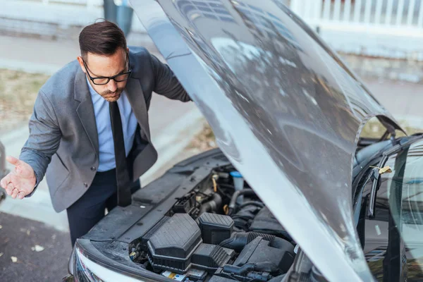 Resmi kıyafetli bir işadamı, kentsel bir arka planda yakalanan mekanik bir sorunla ilgili olarak arabasının kaputunu inceliyor..
