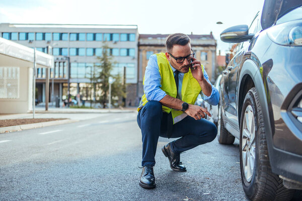 A professionally dressed individual wearing a reflective vest examines a car issue, using their phone for assistance, emphasizing problem-solving, caution, and contemporary urban environment.