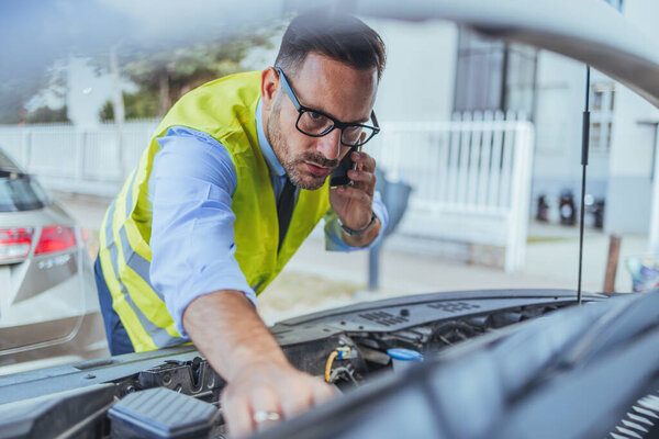 A man wearing a safety vest examines a car engine while making a phone call, indicating a roadside assistance or maintenance scenario.