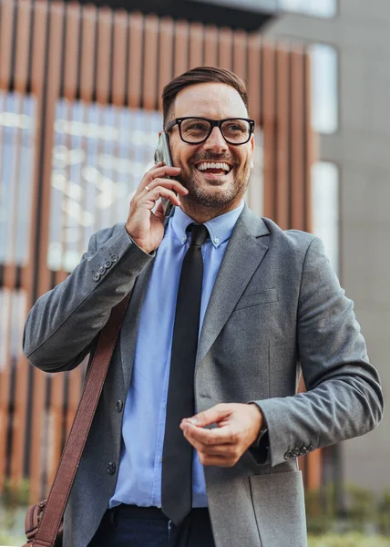 İş kıyafetleri içinde neşeli bir adam telefonuyla konuşuyor, şehir dışında bir yerde güven ve neşe yayıyor. E-posta, başarı, bağlantı ve mesleki başarı kavramlarını çağrıştırıyor.