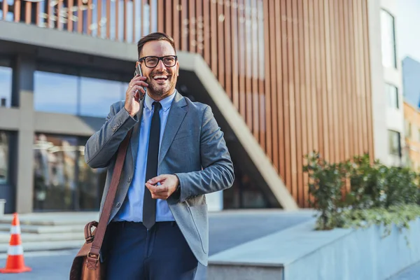 Takım elbiseli ve kravatlı mutlu bir işadamı, omuz çantası tutuyor, telefonuyla çağdaş bir binanın önünde konuşurken gülümsüyor. Görüntü başarıyı, iletişimi ve profesyonel enerjiyi iletir..
