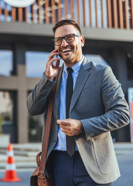 Gri takım elbiseli neşeli bir profesyonel telefonda konuşuyor, pozitiflik ve güven yayıyor. Açık havada, profesyonelliği, iletişimi ve dinamik bir iş ortamını temsil ediyor..
