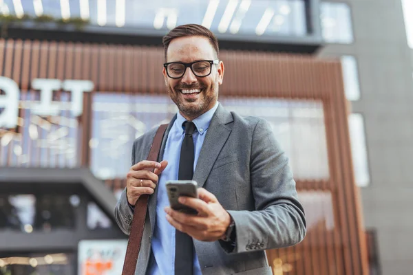 Açık havada akıllı telefon kullanırken gülümseyen takım elbiseli ve gözlüklü profesyonel bir adam. Arka plan modern mimarisi olan modern bir şehir ortamını gösteriyor. İletişim, teknoloji ve kentsel mesleki yaşamı temsil eder.