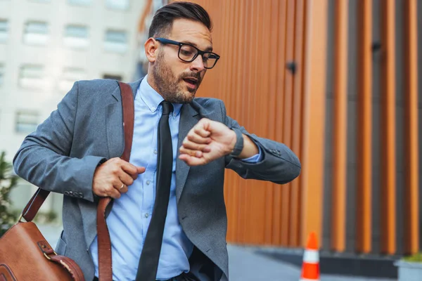 Stresli bir işadamı, modern bir şehir ortamında saatine bakarak bir yere acele eder. Zaman, aciliyet ve yoğun bir şirket hayatı hakkında endişeli görünüyor..