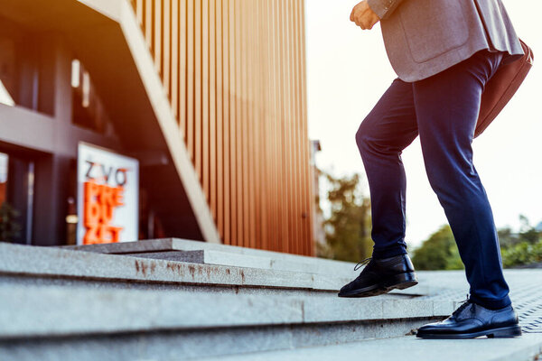 Профессиональный человек в стильной одежде, поднимающийся на конкретные шаги к корпоративному офису в солнечной городской среде, символизирующий прогресс, амбиции и прогрессивный дух бизнес-профессионалов.