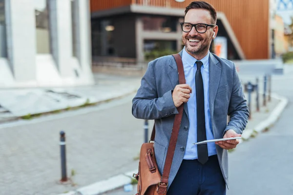 Elinde tablet tutarken gülümseyen profesyonel giyimli bir adam, kendine güven ve profesyonellik yayıyor, şehir ortamında dışarıda duruyor, modern çalışmaları ve olumlu davranışları vurguluyor..
