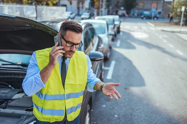 Güvenlik yeleği giyen bir adam yol kenarındaki bozuk arabasının yanında duruyor ve yardım için telefon ediyor. Sahne, kentsel bir ortamda acil durum ve sorun çözmeyi iletiyor.