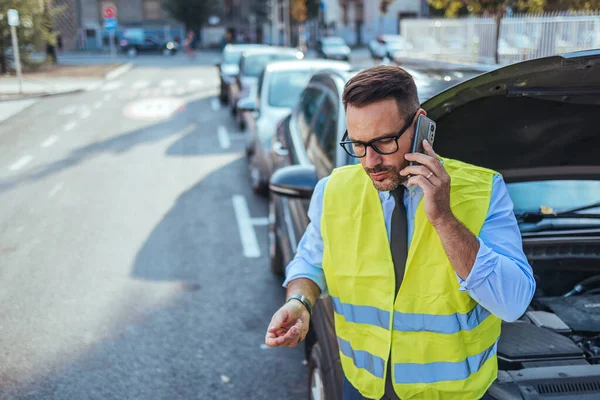 Emniyet yeleği giyen bir adam, şehir sokağındaki bozuk arabasının yanında yol yardımı için telefon ediyor..
