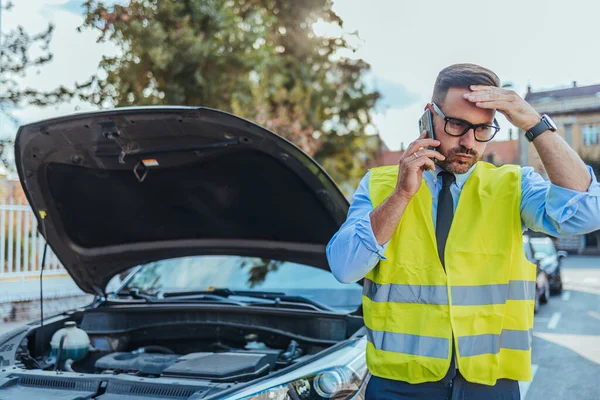 Emniyet yeleği giyen endişeli bir adam, güneşli bir günde bozuk arabasının yanında dururken yol kenarında yardım istiyor..