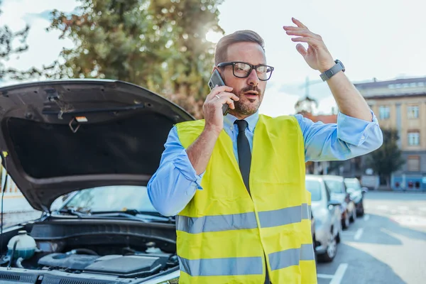Emniyet yeleği giyen hayal kırıklığına uğramış bir adam bozuk bir arabanın yanında duruyor ve cep telefonundan yol yardımı istiyor. Araba işlek bir şehir caddesinde park halinde..