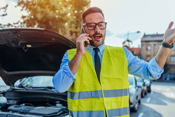 Разочарованный мужчина в спасательном жилете стоит рядом со сломанной машиной, звонит и просит помощи у дороги в солнечный день..