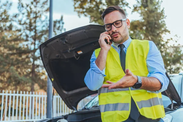 Yansıtıcı güvenlik yeleği giyen bir adam, bozuk bir arabanın yanında acil bir durumu bildiren bir telefon görüşmesi yapar..