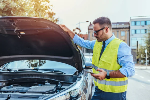 Yansıtıcı güvenlik yeleği giyen bir adam akıllı telefon kullanarak şehir caddesinde bir araba motorunu inceliyor. Olay yeri araç bakımını ve yol yardımını iletiyor.