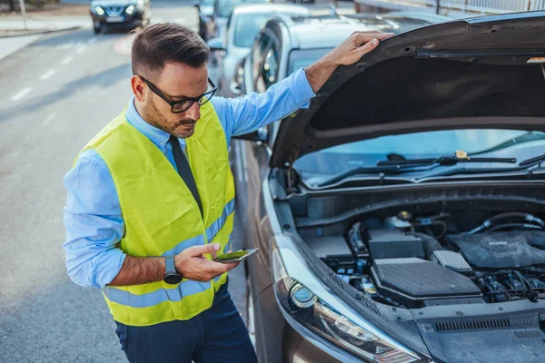 Yansıtıcı yelek giyen bir adam, güneşli bir günde yol kenarındaki cep telefonundan yardım isterken bir araba motoru arızasını inceliyor..