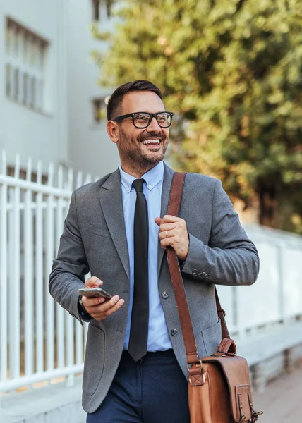 Kendine güvenen bir iş adamı, takım elbiseli ve gözlüklü, elinde akıllı telefon ve deri bir çantayla dışarıda dolaşıyor. Şehir ortamında mutlu ve profesyonel görünüyor..