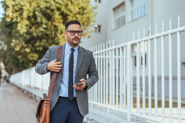 Takım elbiseli bir işadamı kaldırımda yürüyor, deri bir çanta taşıyor, akıllı bir telefon tutuyor, hareket kabiliyetini ve mesleki yaşam tarzını ön plana çıkarıyor..