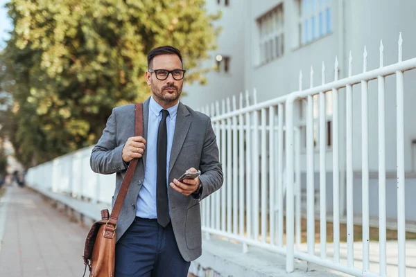 Takım elbiseli kendine güvenen bir işadamı elinde akıllı telefon ve evrak çantasıyla dışarıda dolaşır, profesyonelliği ve modern şehir hayatını simgeler..