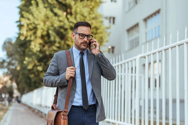Takım elbiseli ve gözlüklü kendine güvenen işadamı akıllı bir telefonla konuşurken dışarıda yürüyor, profesyonellik yayıyor ve işine odaklanıyor. İş, iletişim ve kariyer temaları için ideal.