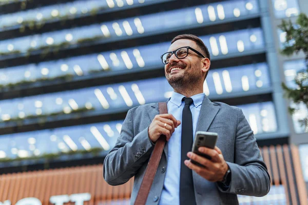 Gülümseyen bir iş adamı arka planda modern bir ofis binası olan akıllı bir telefonla dışarıda duruyor. Başarıyı ve şehir yaşam tarzını simgeleyen güven ve profesyonellik yayıyor..