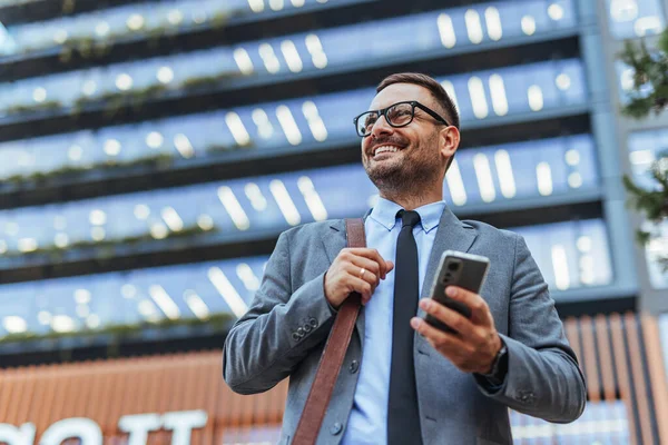 Takım elbiseli ve gözlüklü gülümseyen bir işadamı modern bir ofis binasının önünde dururken elinde akıllı telefon tutuyor. Profesyonel bir şehir ortamında güven anı yakalamak.