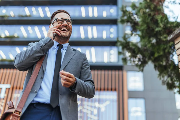 Takım elbiseli gülümseyen işadamı, güneşli bir günde başarı ve iletişimi simgeleyen modern bir ofis binasının önünde telefon görüşmesi yapıyor..