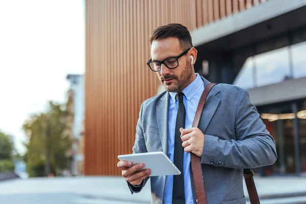 Çağdaş bir ofis binasının önünde dikilirken tablet kullanan takım elbiseli profesyonel bir işadamı iş dünyasındaki teknoloji ve iletişimi aktarıyor..