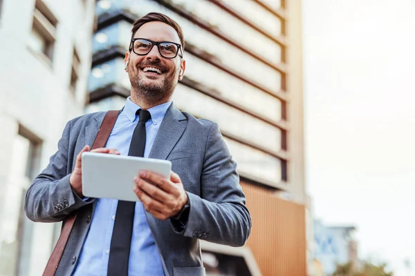 Neşeli bir işadamı şehir ortamında elinde tabletle güvenle dışarıda duruyor. Profesyonel ve başarılı görünüyor, şehrin temiz havasının tadını çıkarıyor..