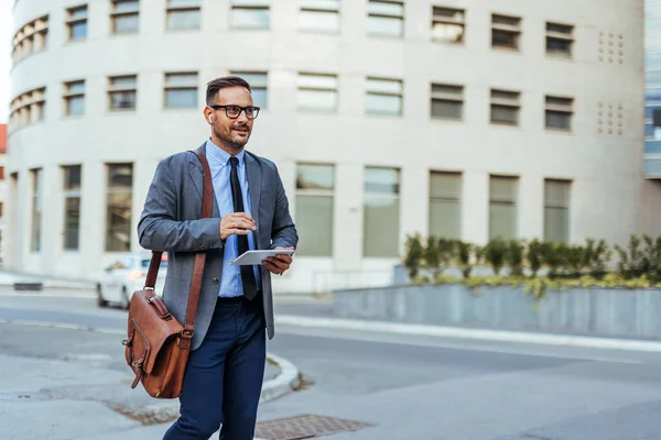 Resmi giyinmiş kendine güvenen bir işadamı elinde tablet ve deri bir çanta ile modern bir ofis binasının önünde yürüyor. İş, şirket ve profesyonel temalar için ideal.