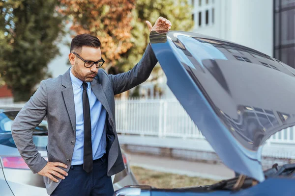 Resmi giyinmiş bir işadamı, şehir caddesinde bozuk bir arabaya sinirlenmiş gibi görünüyor. Sorunu incelerken kaput açık. Beklenmedik arızalar ve mekanik sorunlar kavramı.