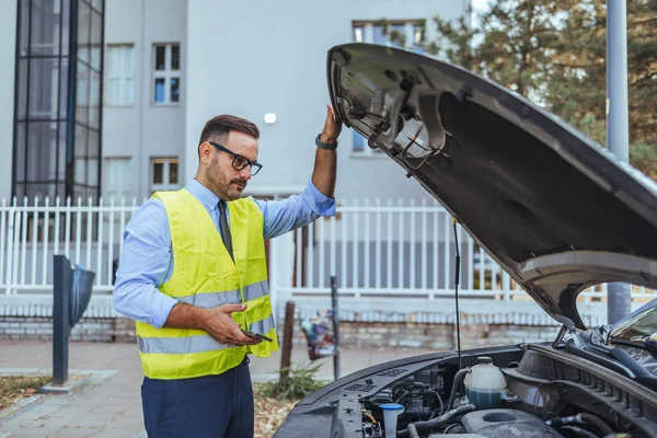 Yansıtıcı yelek giyen bir tamirci açık havada bir arabanın motorunu inceliyor. Şehir ortamında odaklanmış ve profesyonel bir araç denetimi sergiliyor. Otomotiv veya mekanik iş temaları için ideal.