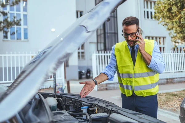 Yüksek görüş mesafesi olan bir adam açık bir araba kaputunun yanında duruyor, telefonla konuşuyor, şehir merkezinde bir araç arızası için yardım istiyor..