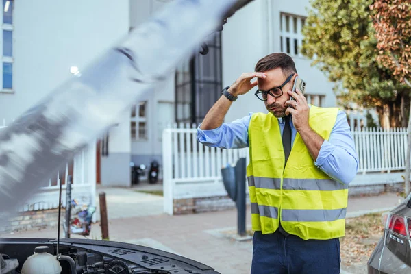 Yansıtıcı yelek giyen endişeli bir adam şehir caddesinde kaputu açık bozuk arabasının yanında bir telefon görüşmesi yapar..