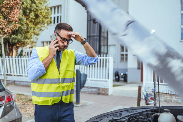 A man wearing a reflective vest calls for assistance with his car broken down on a city street. He looks concerned while seeking help.