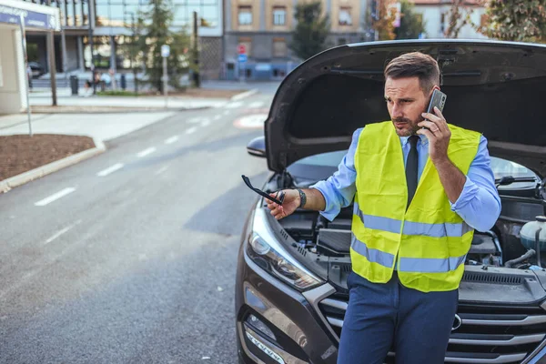 Yansıtıcı yelek giyen bir adam, şehir sokağında bozulmuş bir arabanın yanında yol yardımı ararken telefonda. Endişeli görünüyor ve elinde anahtarlar var..