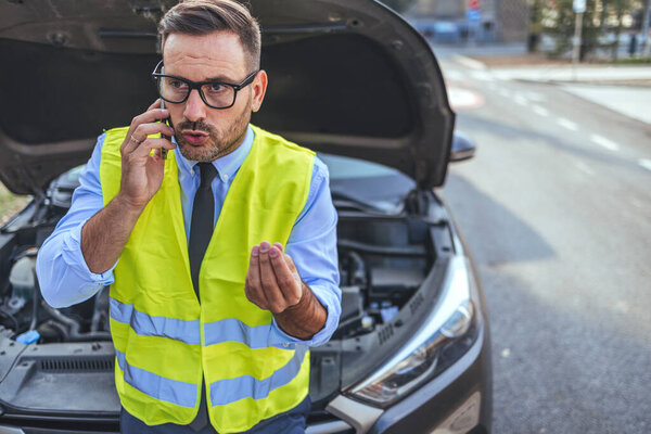 Бизнесмен в спасательном жилете делает телефонный звонок рядом со сломанной машиной. Он выглядит расстроенным, когда жестикулирует, стоя на пустой дороге с открытым капюшоном.