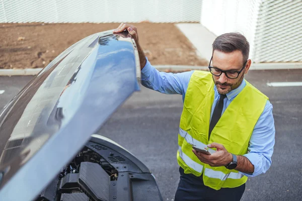 Yüksek görüş açısına sahip yelek giyen bir tamirci akıllı telefonunu kontrol ederken bir araba motorunu kontrol eder. Görüntü, kentsel bir ortamda profesyonellik, teknoloji kullanımı ve araç bakımını iletir.