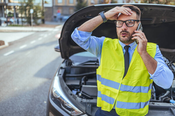 A worried man wearing a yellow safety vest talks on his phone next to his broken-down car. The car hood is up, suggesting engine trouble in an urban setting.