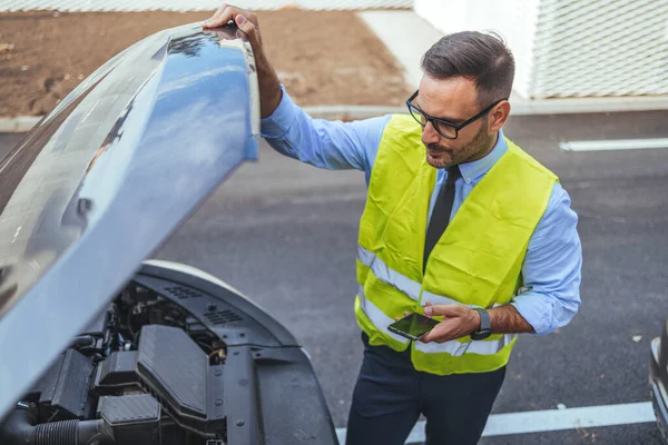 Yansıtıcı güvenlik yeleği giyen bir adam araba motorunu teftiş eder, yol yardımını ve araba bakımını gösterir. Görüntü, yol kenarındaki acil durumlarda sorumluluk ve tedbiri gösteriyor..