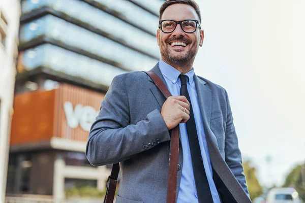 Takım elbiseli neşeli bir işadamı güven içinde şehirde yürür. Gülümsüyor ve bir omuz çantası taşıyor. Şehir ortamında olumlu ve profesyonel bir tavır sergiliyor..