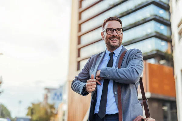 Takım elbiseli ve gözlüklü kendine güvenen bir işadamı pozitif ve başarılı bir şekilde şehirde dolaşıyor. Modern şehir çevresi onun profesyonel görünümünü tamamlıyor..