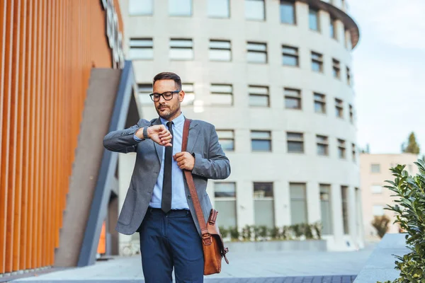 İyi giyimli bir işadamı modern bir ofis binasının önünde durmuş saatini kontrol ediyor. Görüntü, kurumsal bir ortamda profesyonellik, dakiklik ve kentsel yaşam tarzının temalarını aktarır..