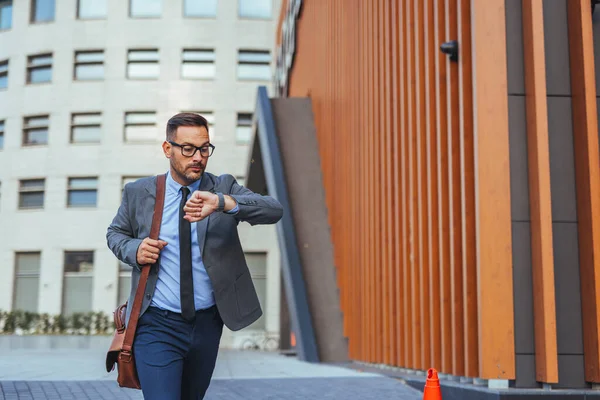 Takım elbiseli odaklanmış bir işadamı modern bir şehir bölgesinde koşuşturup saatini kontrol ediyor. Görüntü, aciliyet, profesyonellik ve hızlı şehir yaşam tarzını yakalıyor..