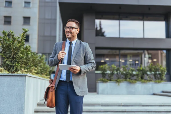 Gülümseyen bir işadamı elinde tabletle bir ofis binasının önünde güvenle duruyor. Profesyonel bir takım elbise giyip, şehir ortamında başarı ve modern iş hayatı yayıyor..