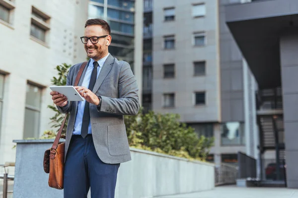 Gülümseyen işadamı, kulaklık takarken tablet kullanarak dışarıda duruyor. Profesyonel olarak giyinmiş, özgüvenini ve modern teknoloji kullanımını ifade ediyor. Kentsel arka plan bir şehir iş ortamı öneriyor.