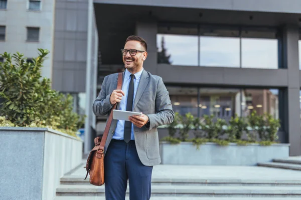 Kendine güvenen bir işadamı modern bir ofis binasının önünde tablet tutarken gülümser. Takım elbise giymiş, profesyonelliği ve başarıyı iletiyor. Yeşillik şehir ortamına biraz doğa katıyor..