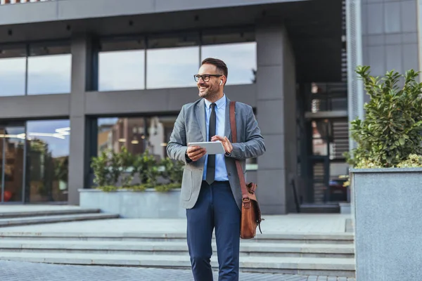 Takım elbiseli gülümseyen işadamı elinde tabletle bir ofis binasının önünde yürüyor. Modern iş, güven ve dijital teknolojiyi temsil ediyor. Çalışmaya ve yeni fırsatlara hazır bir profesyonel..