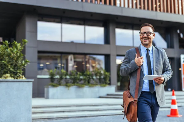 Takım elbiseli neşeli bir işadamı elinde tabletle bir ofis binasının önünde yürüyor. Olumlu ve başarılı bir iş günü için hazır olduğunu gösterir..
