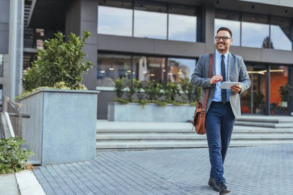 Akıllı takım elbiseli profesyonel bir işadamı tablet taşıyor ve çağdaş bir ofis binasının dışında güvenle yürüyor. Bu görüntü profesyonelliği, güveni ve modern iş yaşamını yansıtır..