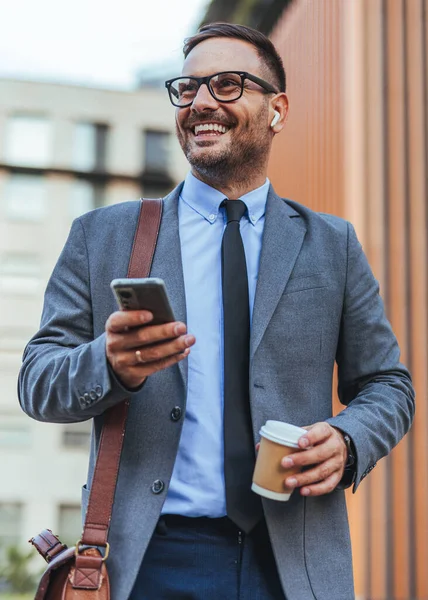 Elinde kahve fincanı ve akıllı telefon tutarken gülümseyen takım elbiseli profesyonel bir adam, kulaklıklarla dışarıda yürüyor. Kentsel bir ortamda başarılı iş, güven ve modern yaşam tarzını temsil eder.